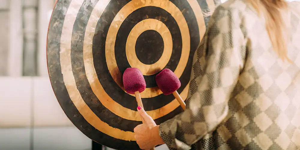 A person holding two purple mallets poised to strike a large, circular gong with concentric black and gold rings.