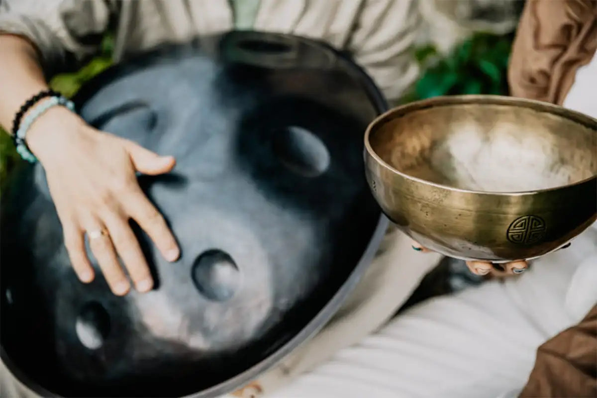 Black handpan with circular sound holes and a brass singing bowl held in hands.