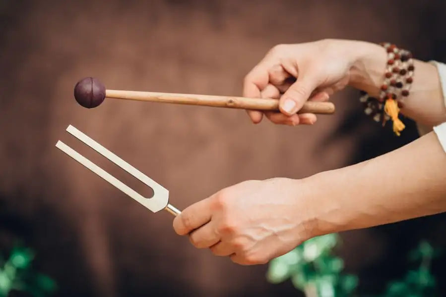 A silver-toned tuning fork with a wooden handle, held by hands adorned with beaded bracelets.